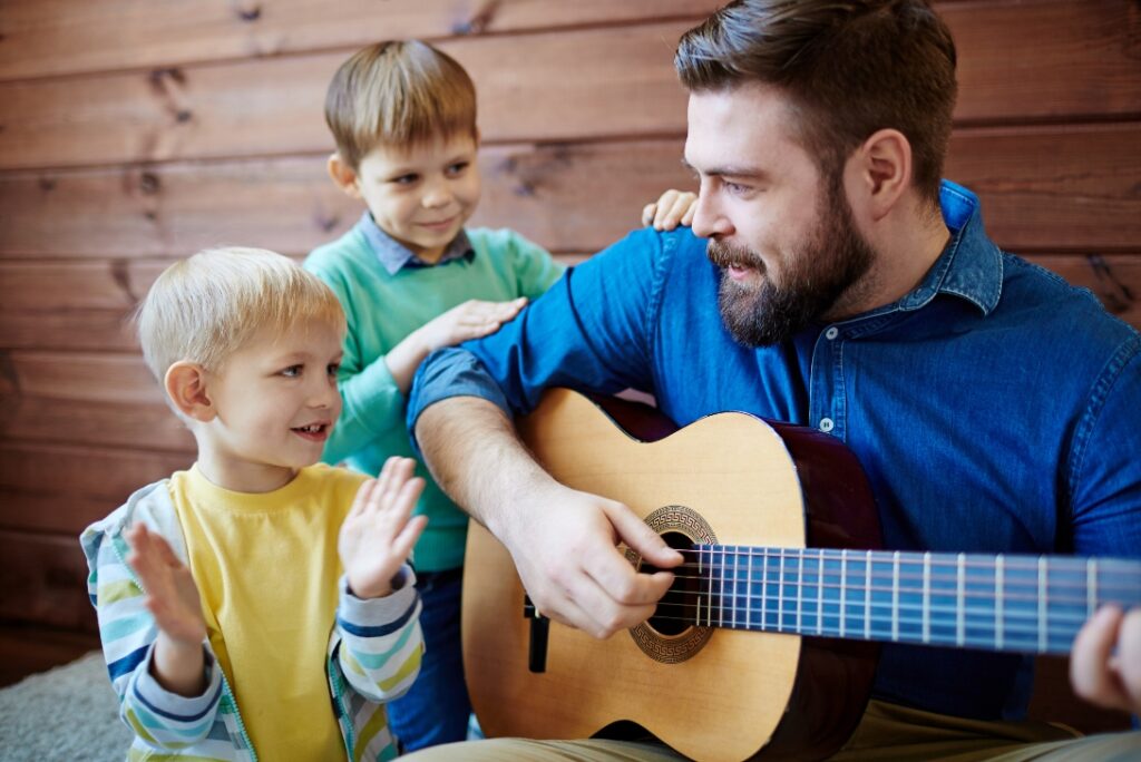 Ein Mann spielt zwei Jungen etwas auf einer Gitarre vor.