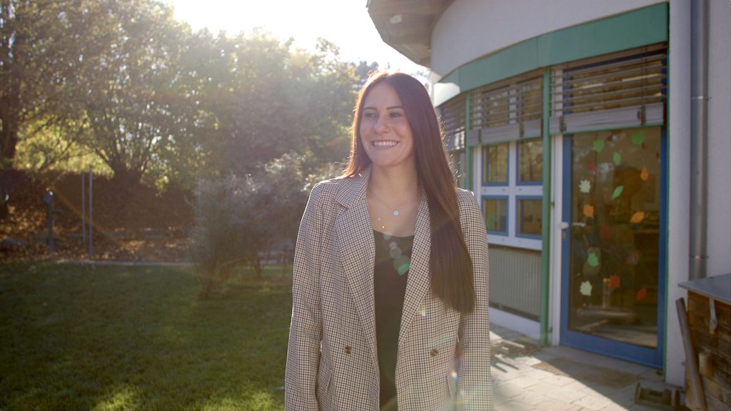Eine junge Frau steht draußen vor einem Kindergarten und guckt lächelnd in die Ferne. 
