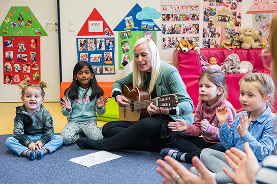 Eine Frau spielt mehreren Kindern etwas auf einer Gitarre vor. Sie sitzen zusammen auf dem Boden und die Kinder klatschen zur Musik.