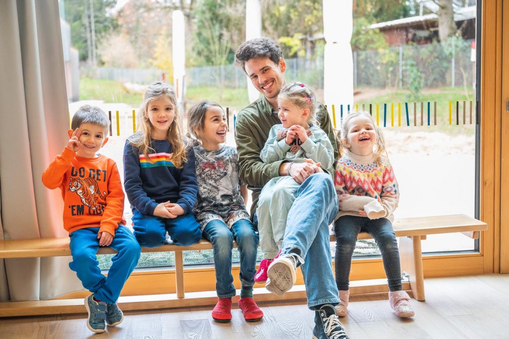 Ein junger Mann sitzt mit 5 Kindern auf einer Bank, alle lachen in die Kamera. 