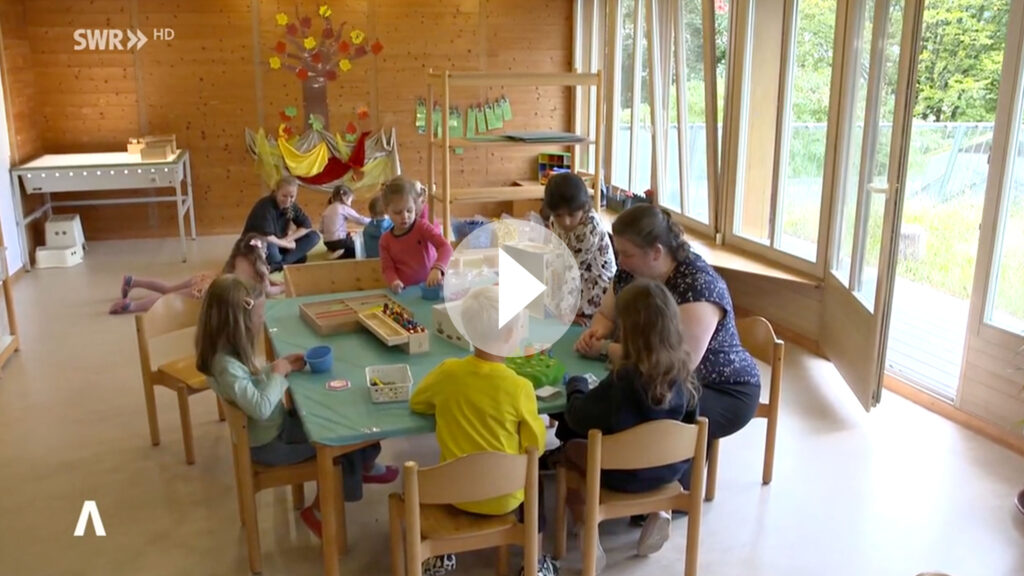 Man sieht ein Spielzimmer einer Kita, eine Erzieherin sitzt mit einigen Kindern zusammen an einem Tisch. 