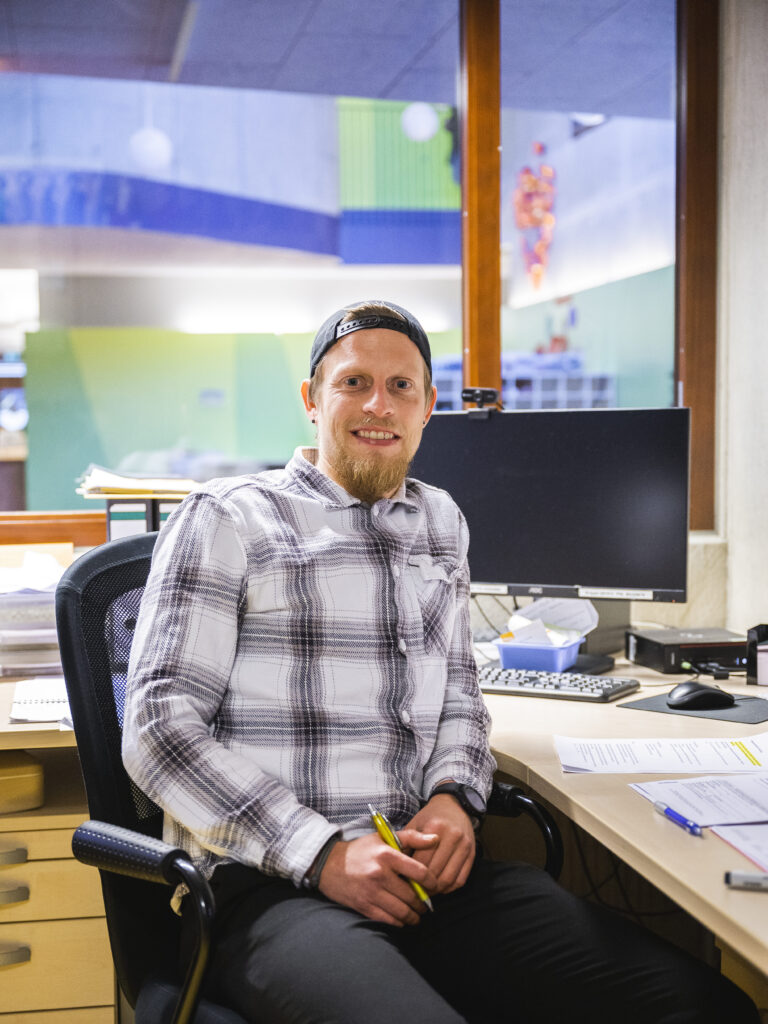 Der Kindergärtner sitzt an seinem Schreibtisch und lächelt in die Kamera.