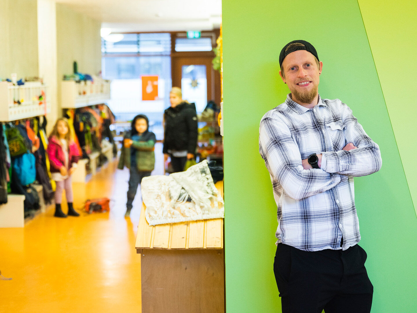 Ein Mann lehnt an einer Wand während sich im Hintergrund Kinder ihre Jacken anziehen.